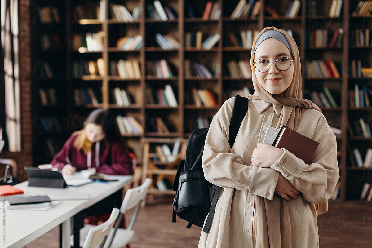 Positive Muslim female student standing with book in college library