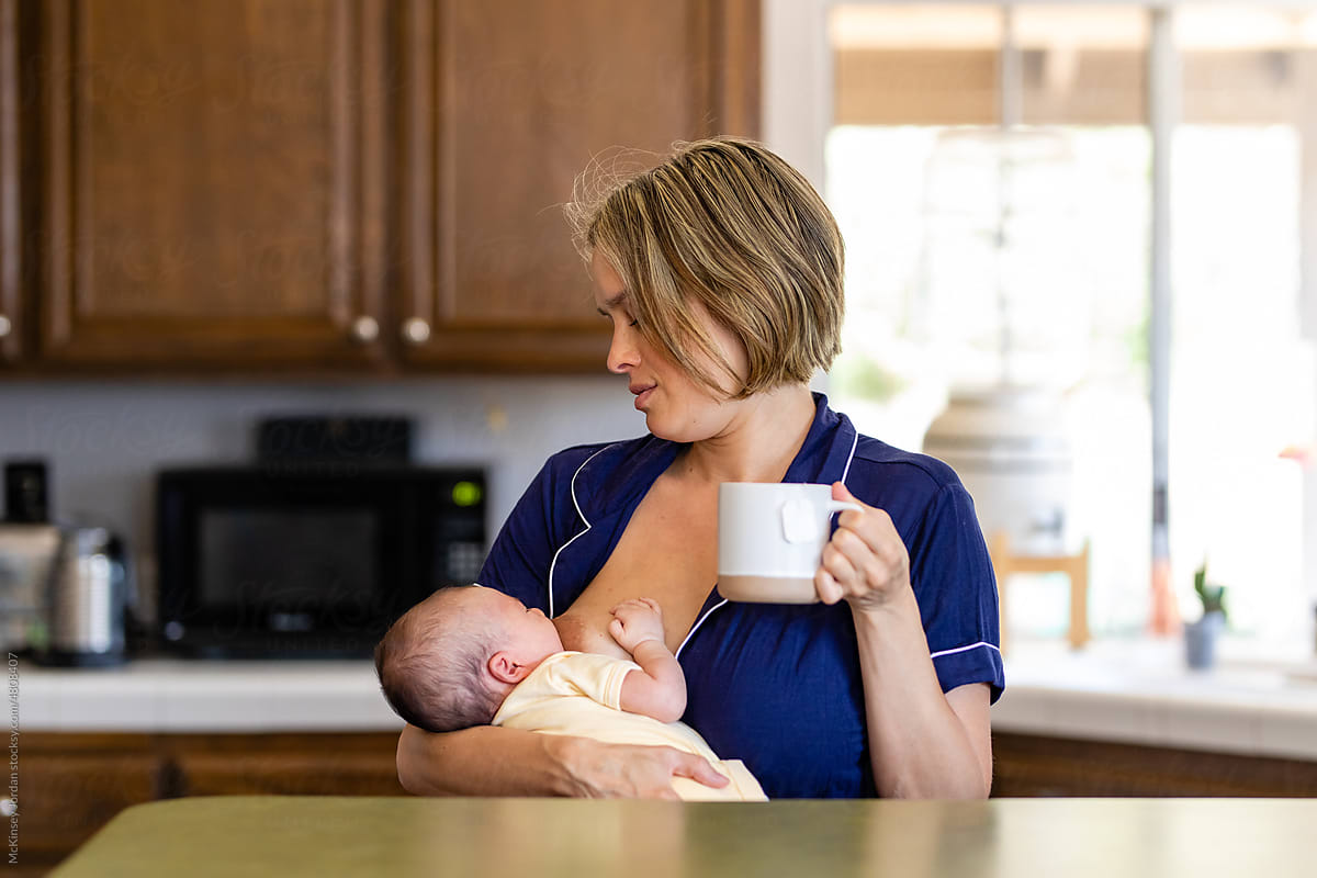 Woman Enjoys A Cup Of Tea While Nursing Her Newborn In The Kitchen