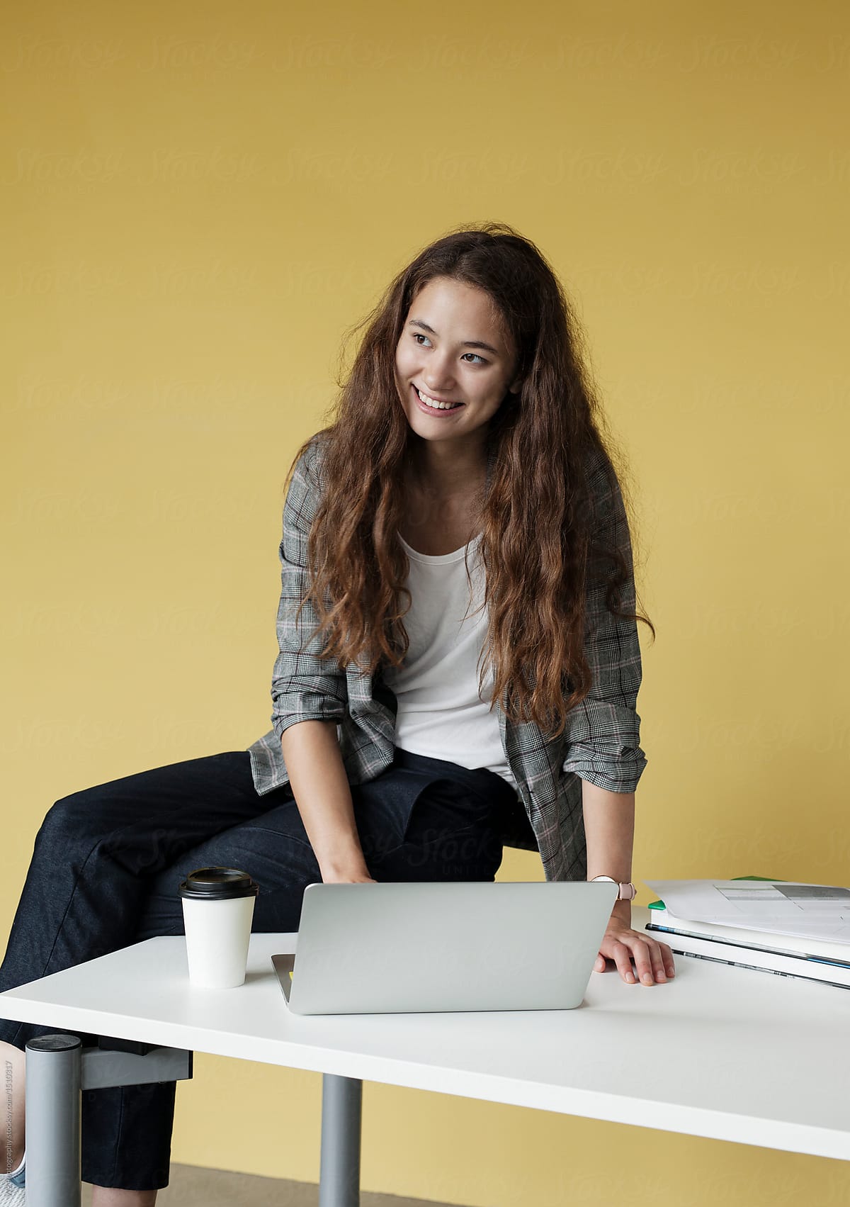 Smiling student sitting on her school desk against a yellow wall.