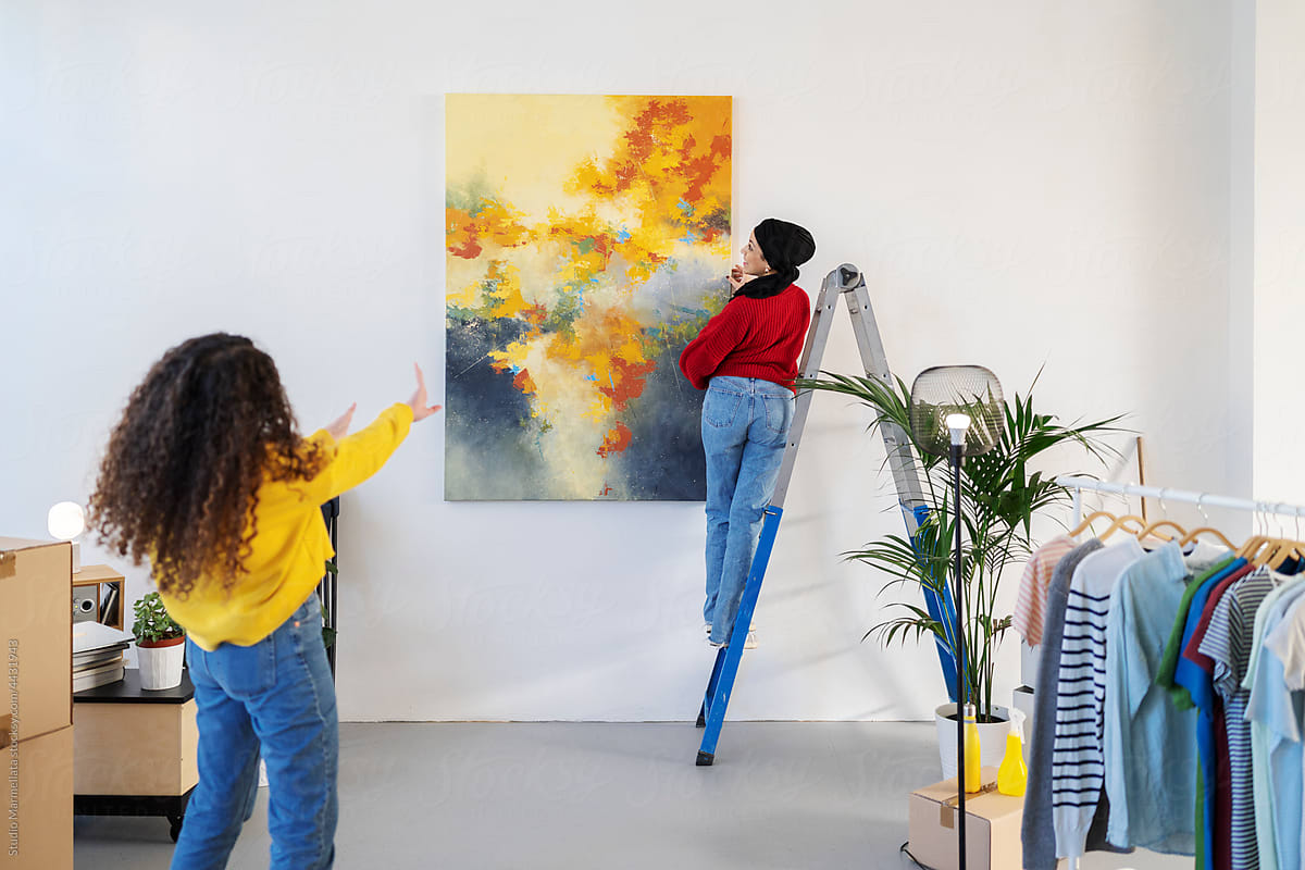 Diverse women hanging painting on wall in house