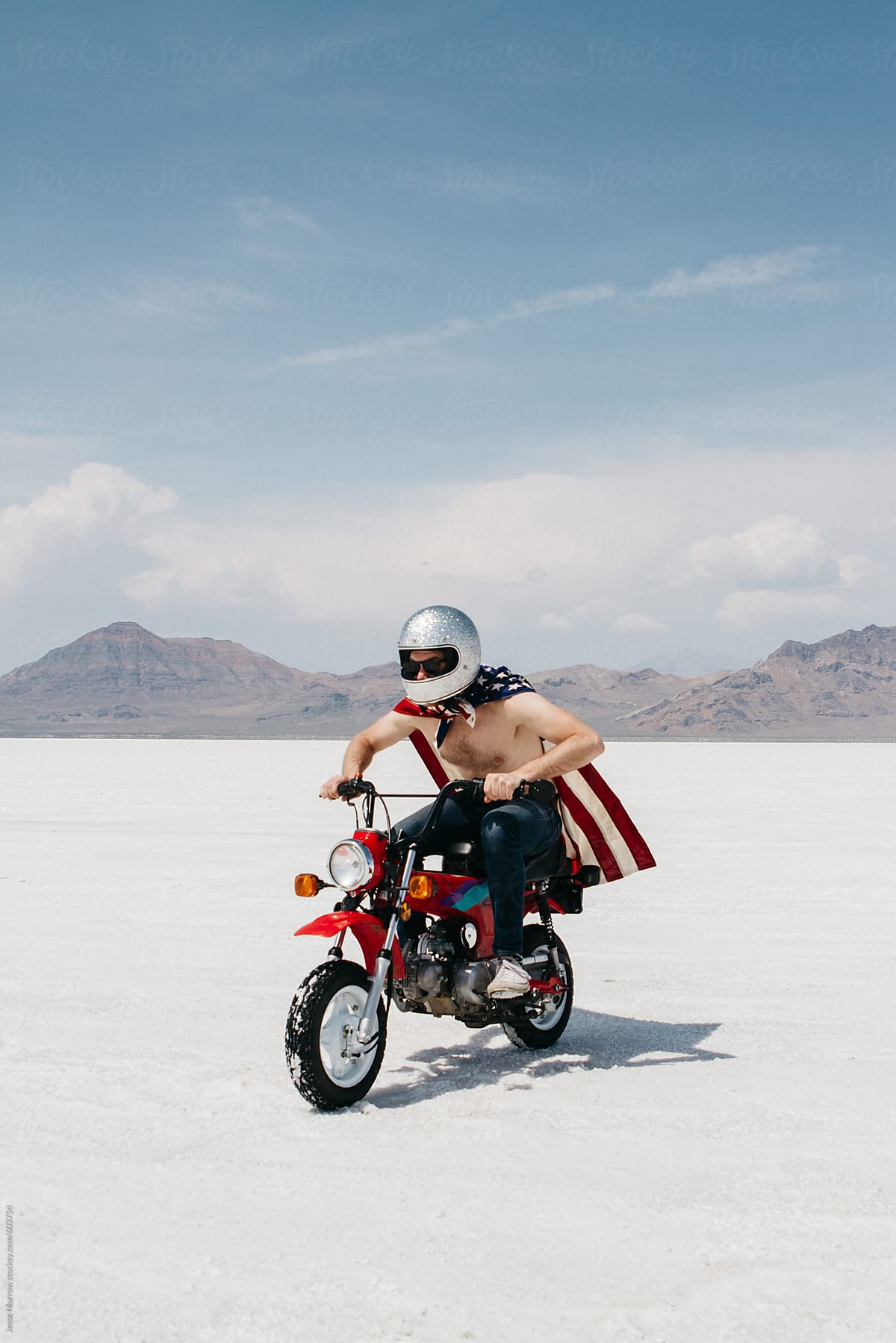 young man with no shirt on wearing american flag while riding small motorcycle in desert salt flats
