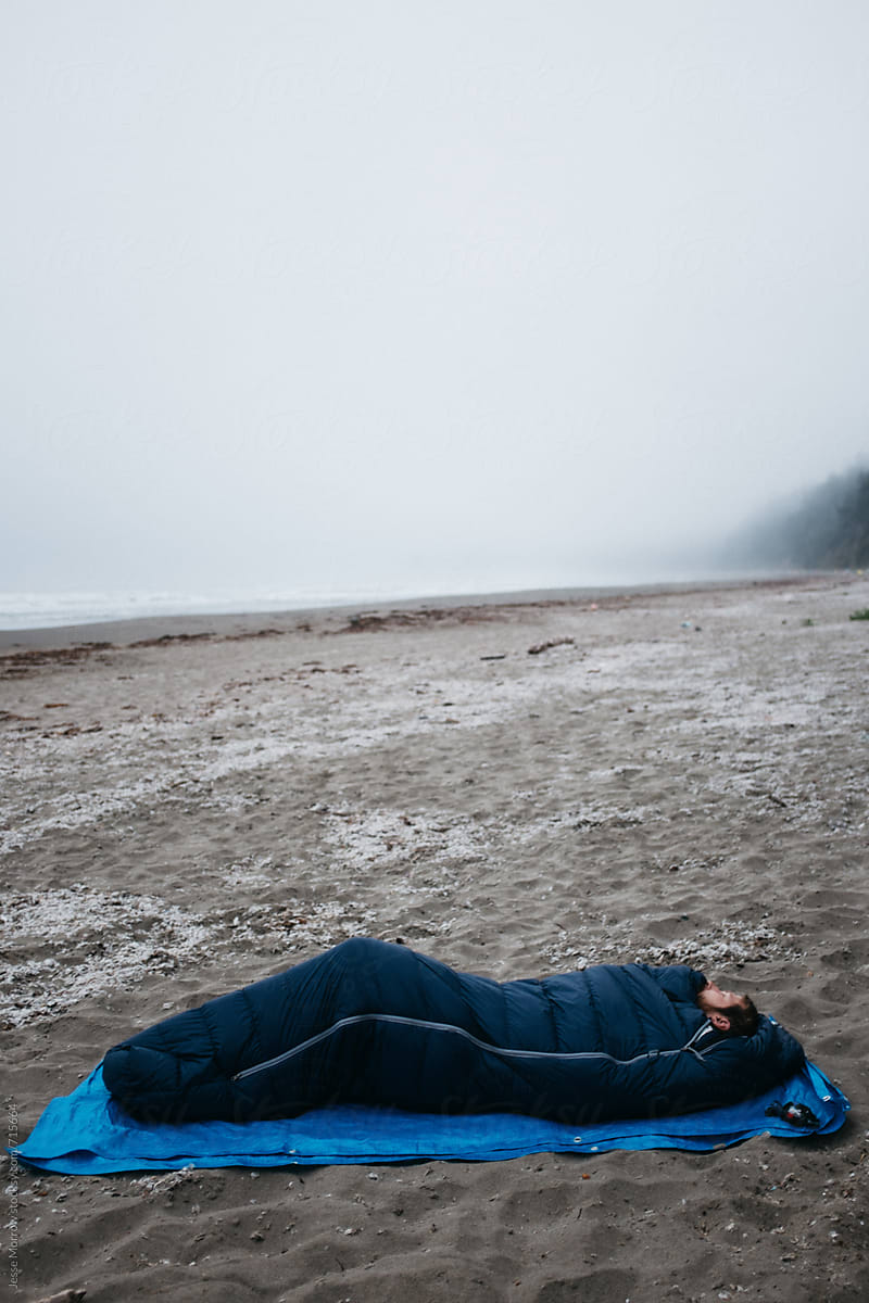 young male sleeping alone on beach in sleeping bag