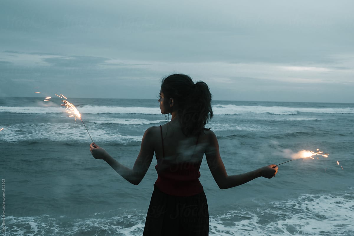 young fashion woman stand on the ocean coast with fireworks sparkles in hands with sea view