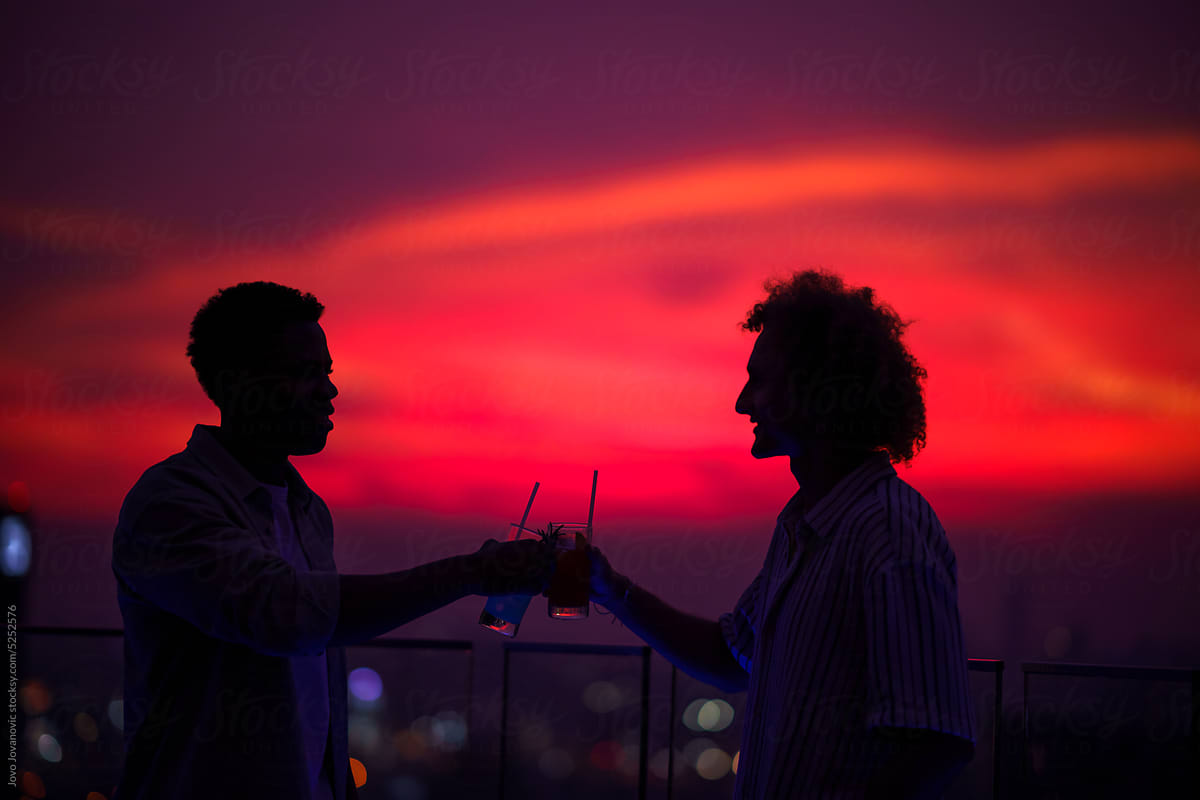 Silhouette of two friends toasting drinks glass on rooftop at sunset