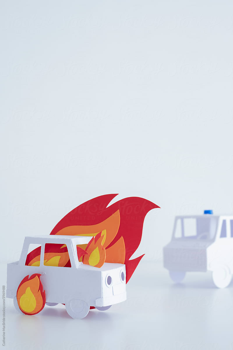 A paper craft car on fire with ambulance in  background.
