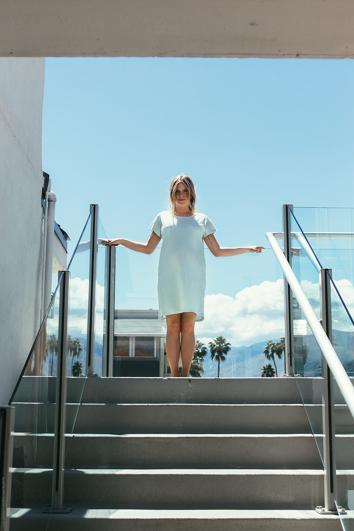 Young Pregnant Woman In Light Blue Dress Standing At Top Of Stairs On Sunny Day