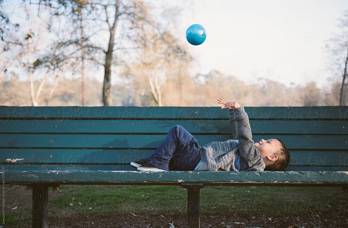Boy lying down on park bench throwing his ball