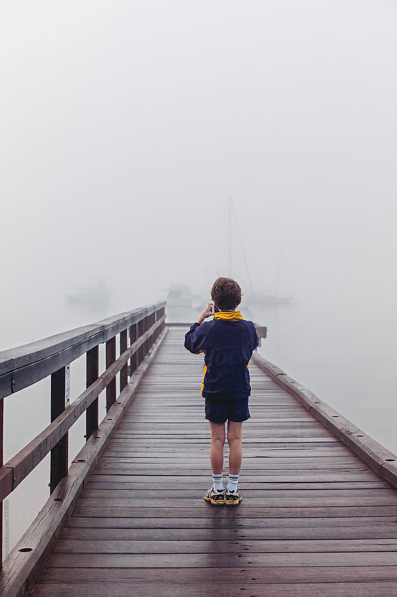 Boy taking photos on a jetty over a river with moored yachts just visible in dense fog
