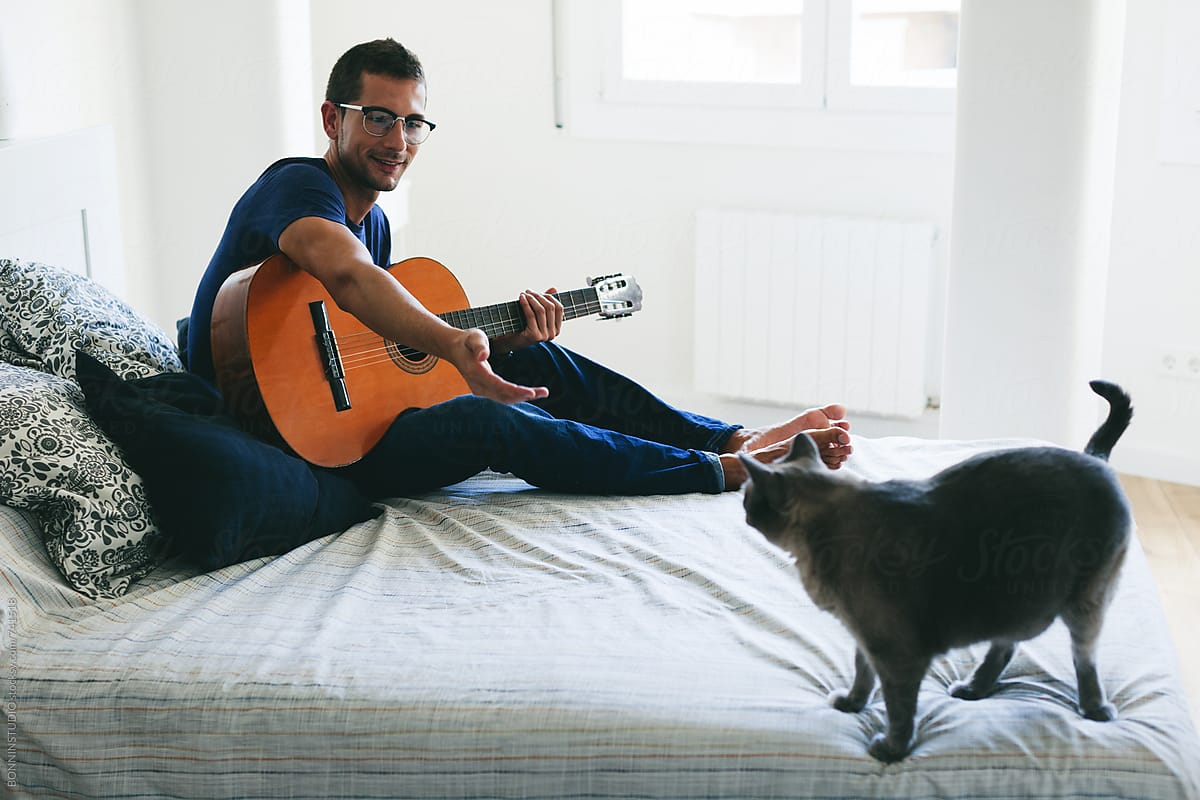 Man playing guitar with his siamese cat on bedroom.
