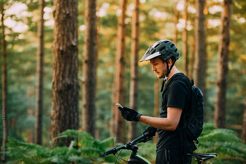 A male bike rider checks his phone in the forest, surrounded by ferns and tree trunks