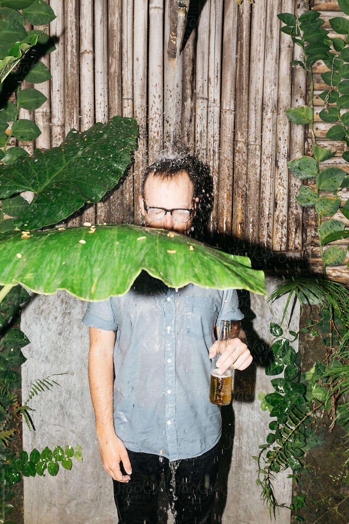 Young man drinks beer fully clothed in outdoor wild shower with large plants