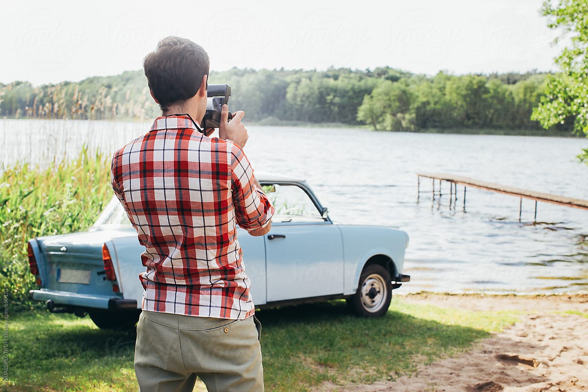 Young Man Taking Polaroid Photo of Vintage Car Parked by Lake