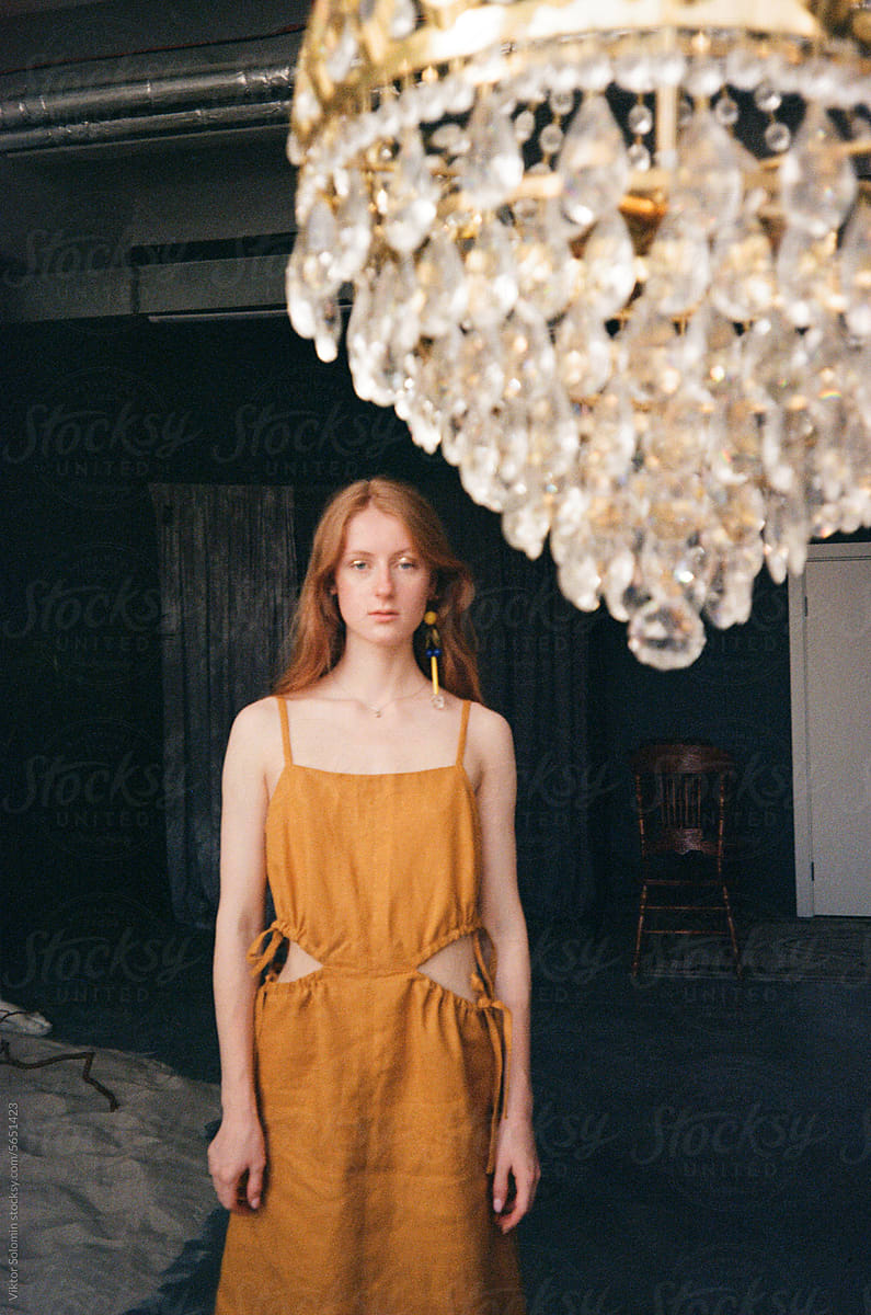 Elegant ginger woman in dress standing in dark room near chandelier