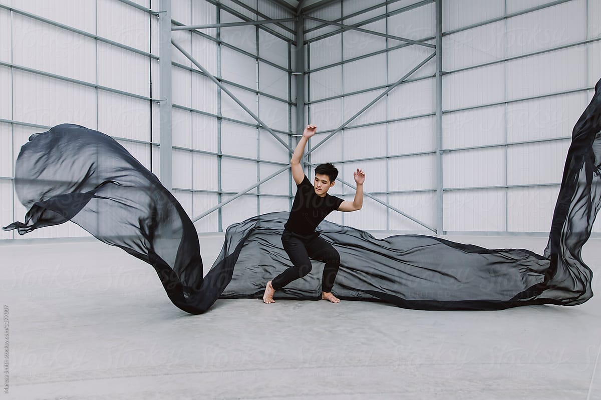 An asian male dancer in a warehouse bracing himself as silk fabric falls around him