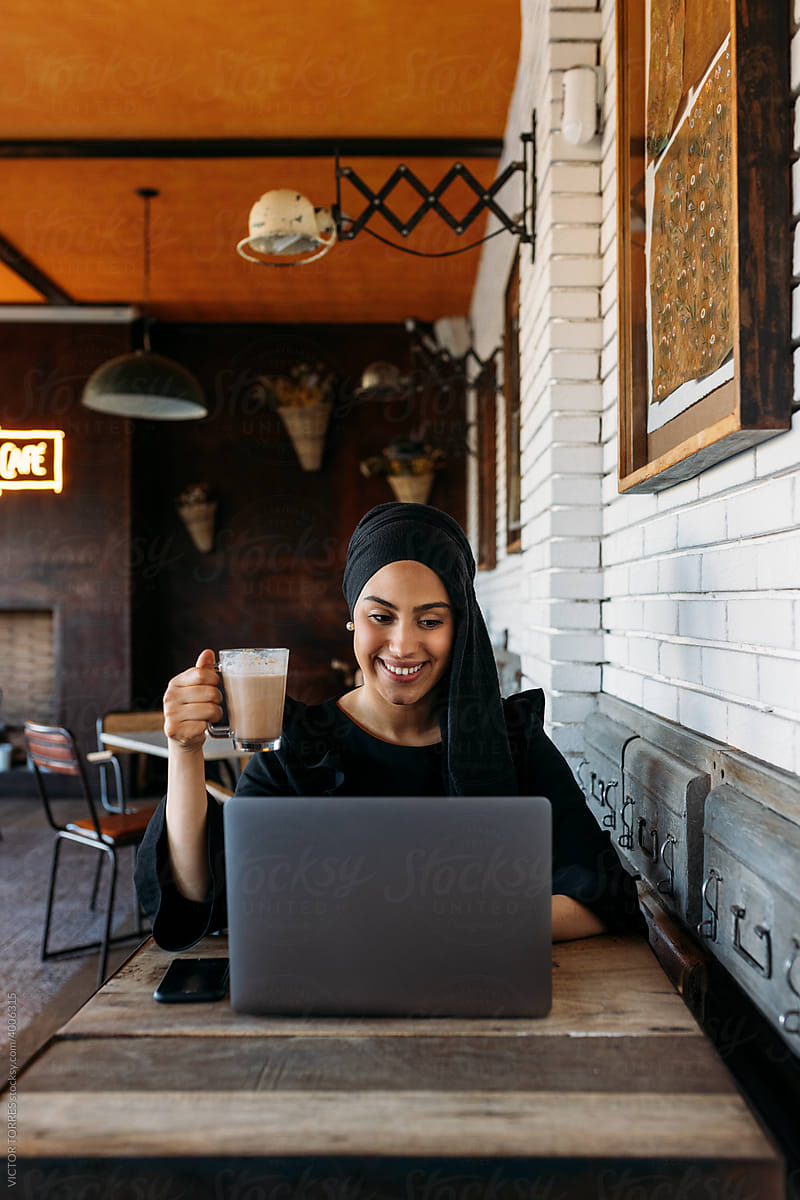 Cheerful stylish Muslim woman using laptop in cafe