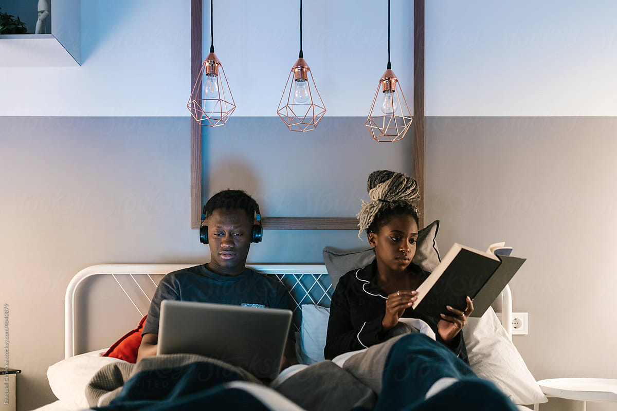 Man using laptop in bed with girlfriend reading book