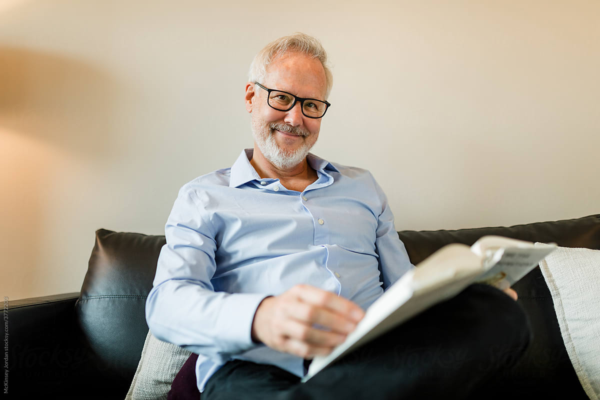 Man Wearing Glasses and Reading on His Couch Smiles at the Camera