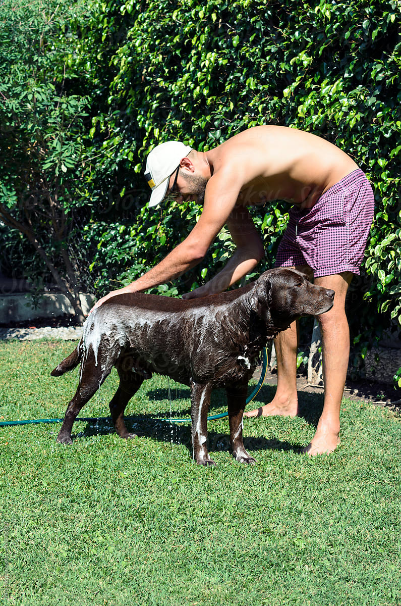 Young man in swimsuit and baseball hat washing his dog