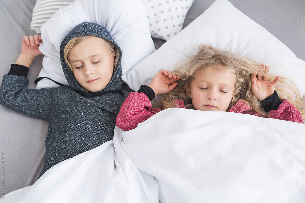 Little girl with white curly hair in red pajamas and a little boy with blond hair in a gray pajamas sleeping in bed.