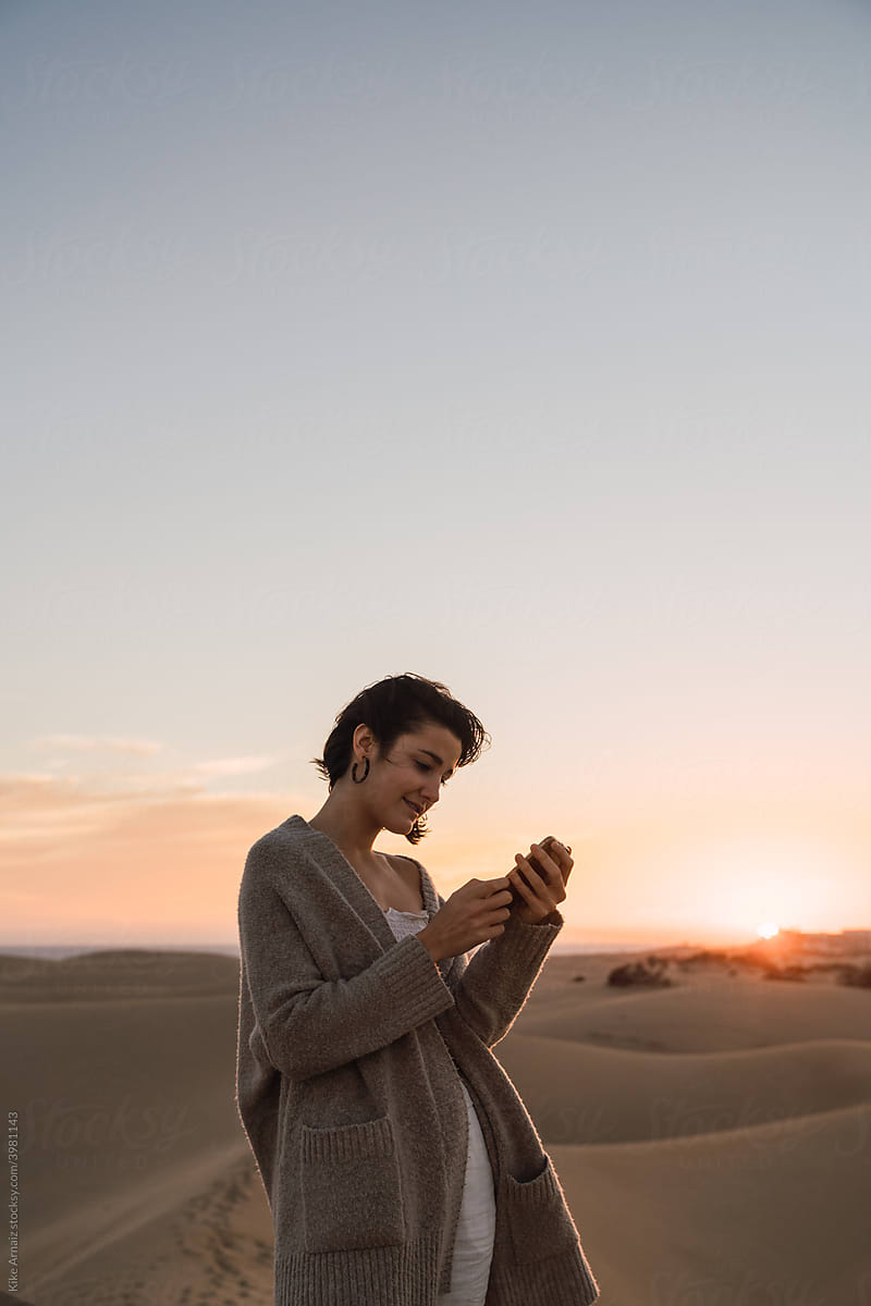 Woman standing with smartphone on sand land and blue sky at sunset