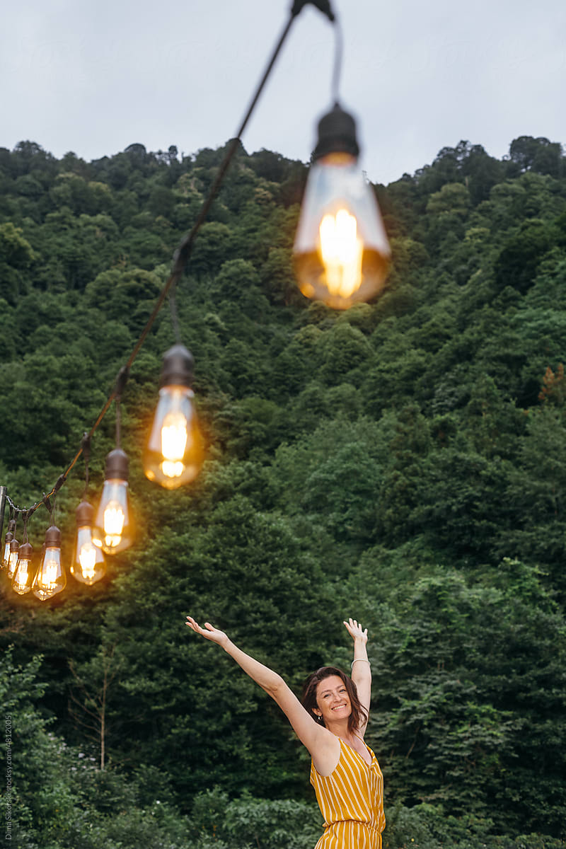 Happy girl with raised hands near light bulbs and forest mountains