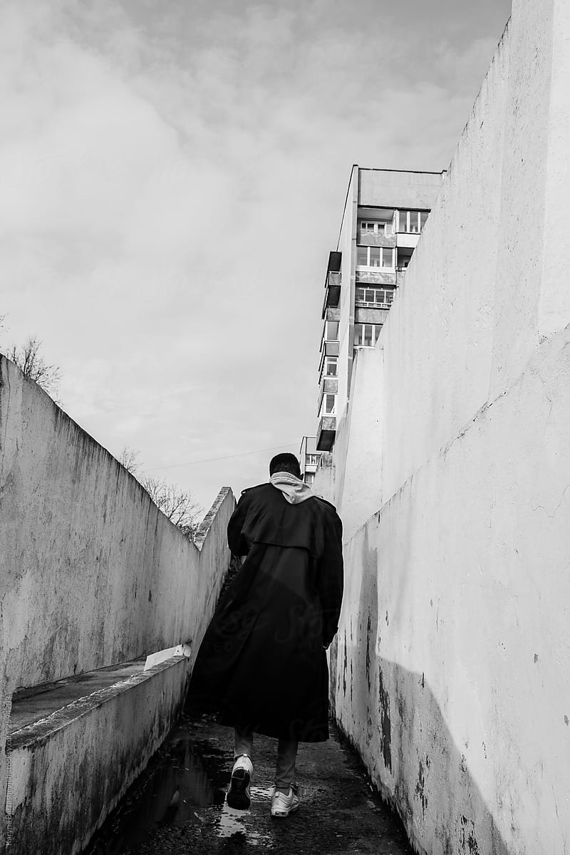 black and white photo of a stylish alien guy walking along the aisles of old buildings in a long coat