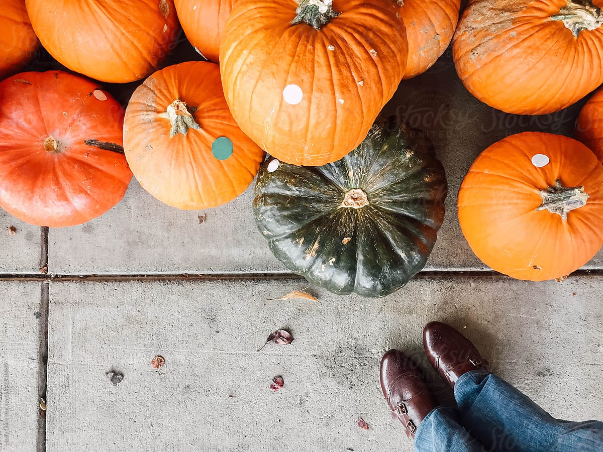 Orange and green pumpkins sitting on concrete sidewalk with denim legs and leather shoes.