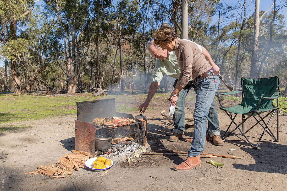 Grey nomad couple tending meat, vegetables and fruit cooking on grill at campfire